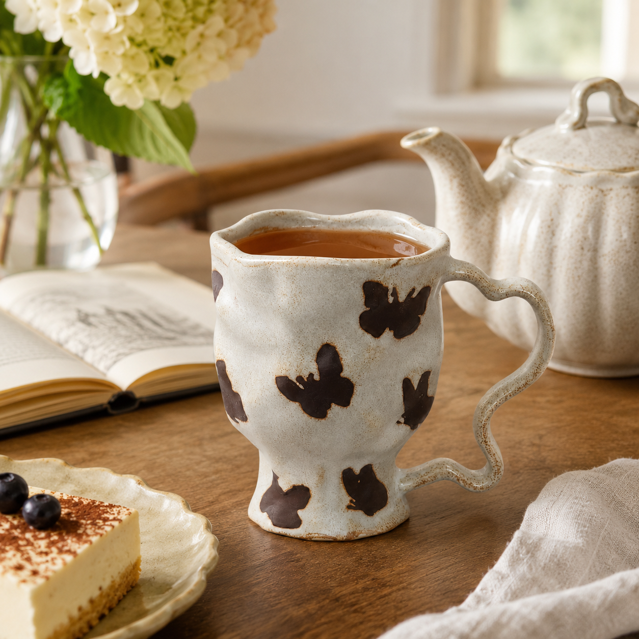 Butterfly ceramic mug filled with tea beside an open book, teapot, and dessert